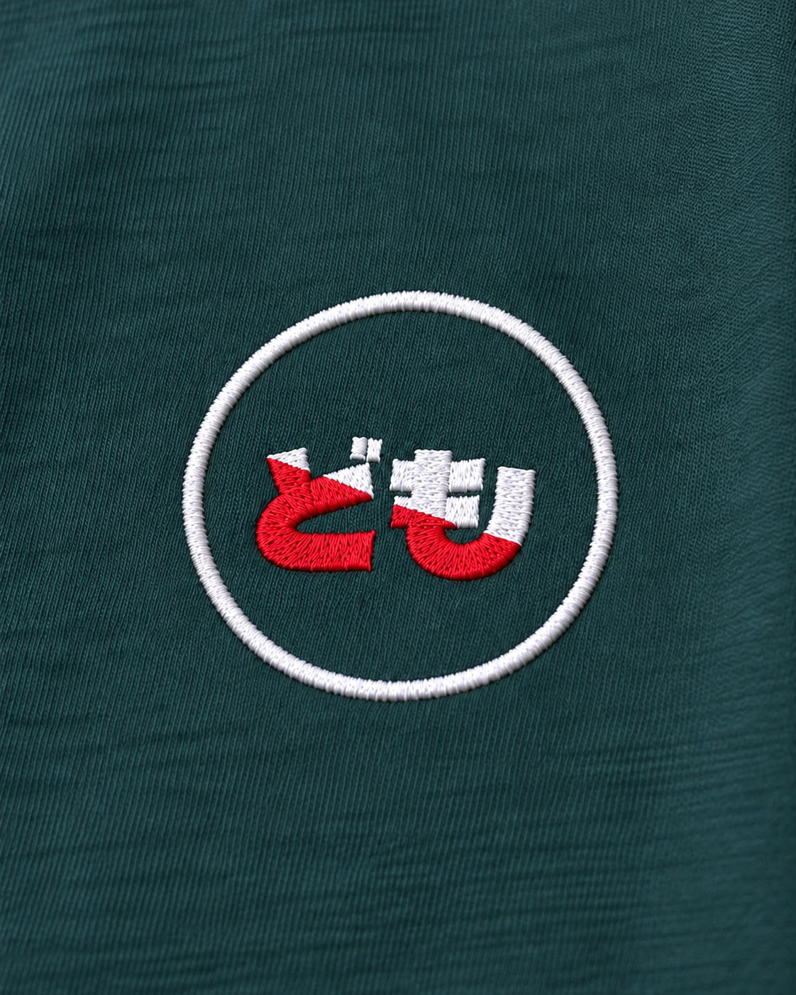 Kata Tee Ink — close-up of the original embroidered Domo chest logo, showing red and white stitching on green cotton
